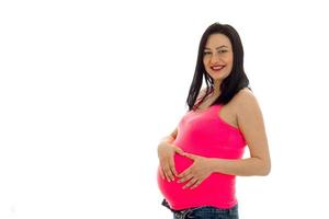 Studioporträt einer jungen schwangeren brünetten Frau in rosa Hemd, die ihren Bauch berührt und vor der Kamera lächelt, isoliert auf weißem Hintergrund foto