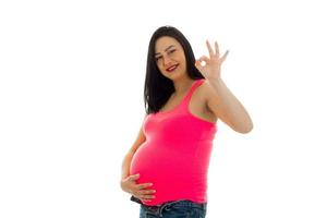 Studioporträt einer jungen schwangeren brünetten Frau in rosa Hemd, die ihren Bauch berührt und vor der Kamera isoliert auf weißem Hintergrund ok zeigt foto