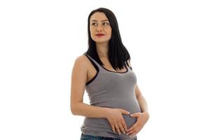 Studioporträt einer jungen schwangeren brünetten Frau im Hemd, die ihren Bauch berührt und zur Seite schaut, isoliert auf weißem Hintergrund foto