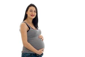 Studioporträt einer jungen schwangeren brünetten Frau im Hemd, die ihren Bauch berührt und isoliert auf weißem Hintergrund in die Kamera schaut foto