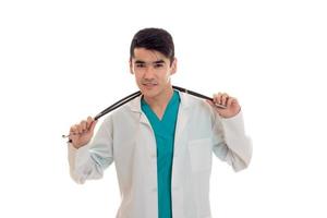 Porträt eines jungen, schönen, brünetten Mannes Arztes in weißer Uniform mit Stethoskop, der isoliert auf leerem Hintergrund in die Kamera blickt foto