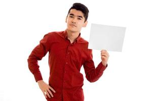 youn attraktives brünettes männliches Modell im roten Hemd, das mit leerem Plakat in seinen Händen aufwirft und die Kamera lokalisiert auf weißem Hintergrund betrachtet foto