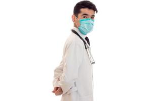Arzt in einem weißen Laborkittel steht seitlich mit einer Maske auf seinem Gesicht isoliert auf weißem Hintergrund foto