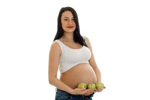 fröhliche schwangere Frau des Brunette, die mit grünem Apfel in ihren Händen lokalisiert auf weißem Hintergrund aufwirft foto