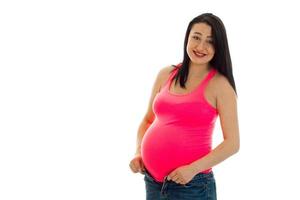 Lustiges schwangeres Mädchen mit dunklem Haar, das ihren dicken Bauch berührt und isoliert auf weißem Hintergrund in die Kamera schaut foto