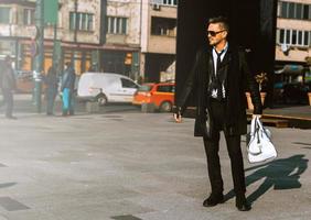 junges erwachsenes männliches Model im Anzug mit Lederjacke, Sonnenbrille und weißer Tasche, die wegschaut foto