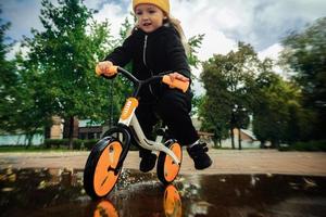 Cutie kleines Mädchen fährt auf Fahrradcutie kleines Mädchen fährt durch die Pfützen auf dem Fahrrad im Herbstpark im Park foto