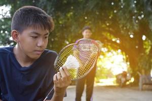 junger asiatischer junge hält traurig gebrochenen rahmen-badmintonschläger in der hand, während er mit seinem freund außerhalb des hauses badminton spielt, badminton-konzept im freien, weicher und selektiver fokus. foto