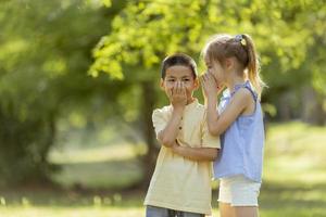 Kaukasisches Mädchen, das dem asiatischen Jungen im Park ein Geheimnis erzählt foto