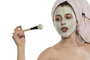 Schöne junge Frau mit einem um den Kopf gewickelten Handtuch, das im Badezimmer eine Gesichtsmaske aus Ton anwendet foto