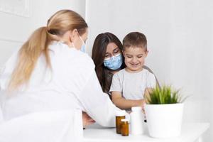 Junge brünette Frau mit kleinem Jungen, die sich im Kinderarztbüro beraten lässt. arzt, kind und mutter tragen während des ausbruchs von coronavirus und grippe gesichtsmasken. Virus Schutz. Covid 19. foto