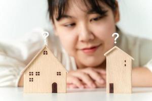 Die Frau entscheidet sich für ein Haus mit einem kleinen Holzhausmodell mit Fragezeichen und plant, eine Immobilie zu kaufen. wählen Sie, was das Beste ist. Konzepte für Wohnungsbaudarlehen. foto