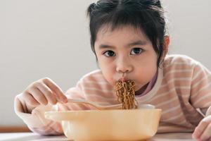 asiatische Babymädchen genießen glücklich mit Bestecklöffel und Gabel, die köstliche Nudeln in der Küche am Esstisch essen. Fröhliches asiatisches Mädchen übt, alleine am Esstisch zu essen. Babynahrungskonzept foto