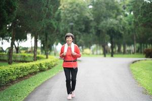 athletische asiatische Seniorin der 60er Jahre lächelt und joggt. Schöne ältere Asiatin, die an einem sonnigen Tag im Park läuft. Läuferin, die beim Joggen Musik hört. foto