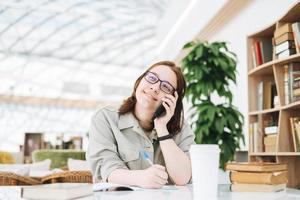 Junge brünette Teenager-Studentin in Brille mit Handy, die an einem öffentlichen Ort in der grünen modernen Bibliothek arbeitet foto