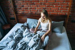 Erwachsene Frau vierzig Jahre alt mit blonden langen Haaren im Schlafanzug mit Handy auf dem Bett im Loft-Zimmer zu Hause foto