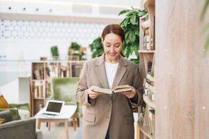 erwachsene lächelnde brünette geschäftsfrau vierzig jahre mit langen haaren in stylischem beige anzug und jeans an öffentlichem ort, grünes open space office, coworking. freundlicher lehrer oder mentor mit buch in der bibliothek foto