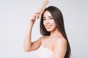 Schönheitsporträt der glücklich lächelnden asiatischen Frau mit dem dunklen langen Haar, das hölzernen Kamm auf dem weißen Hintergrund lokalisiert kämmt foto