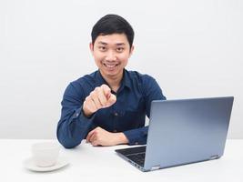 asiatischer mann zeigt mit dem finger auf dich, sitzt mit laptop und kaffeetasse am tisch foto