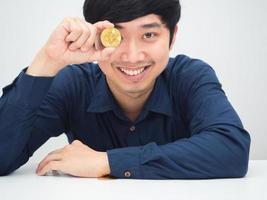 asiatischer mann fröhlich lächelndes gesicht schließt sein auge durch gold bitcoin, das am schreibtisch sitzt foto