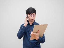 asiatischer Mann mit Brille, der mit dem Handy spricht und sich ernst fühlt, wenn er die Dokumententafel in seinem Handweißhintergrund betrachtet foto