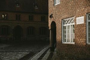 ecke eines alten backsteinhauses mit der unterführung, die von der steinsäule gestützt wird, die von der nachmittagssonne erleuchtet wird, mit der nassen granitstraße herum im ältesten dorf dänemarks rib foto