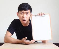 junger asiatischer mann ernstes und verwirrtes gesicht, das weißes papier auf klemmbrett zeigt, sitzt am schreibtisch auf weißem isoliertem, geschäftsarbeitskonzept foto