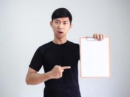 junger asiatischer mann schwarzes hemd zeigen finger auf holz zwischenablage leeres dokument in der hand ernstes gesicht auf weiß isoliert foto
