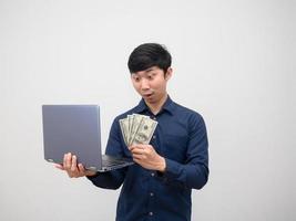 asiatischer mann sieht geld in seiner hand und hält einen laptop, der sich auf weißem hintergrund erstaunt fühlt foto
