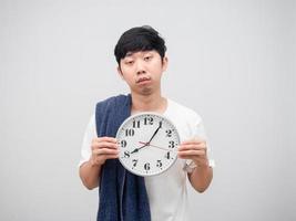 asiatischer mann mit handtuch, das verschlafenes gesicht der uhr auf weißem hintergrund hält foto