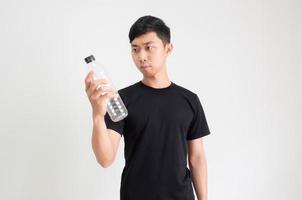 junger asiatischer mann, schwarzes hemd, blick auf plastikflasche in der hand und verwirrtes gesicht auf weiß isoliert foto