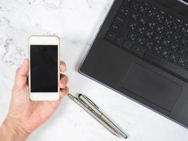 Draufsicht Arbeitsbereich Hand mit Handy mit silbernem Stift und weißem Marmorschreibtisch des Laptops foto