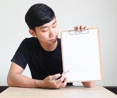 junger asiatischer mann schwarzes hemd fröhlich sitzen am schreibtisch und zeigen zwischenablage zeigen den stift auf leeres dokument, geschäftskonzept foto