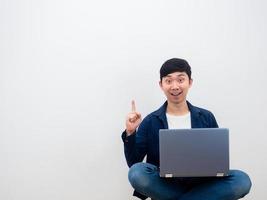 asiatischer mann mit dem laptop, der auf dem stuhl sitzt, bekommt die idee, den finger auf weißen wandhintergrund zu zeigen foto