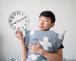 junger mann wacht auf, umarmt kissen und schaut auf die uhr in der hand, fühlt sich gelangweilt im gesicht auf weiß isoliert, wacht spät auf foto