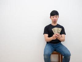 asiatischer mann, der sich mit geld in seiner hand erstaunt fühlt, sagen wow, sitzend auf weißem hintergrund des stuhls foto