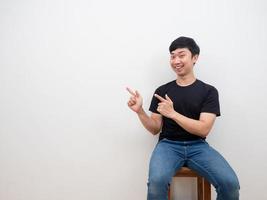 asiatischer mann, der auf stuhl mit lächelngesicht sitzt, fühlt sich glücklich und fröhlich, zeigt mit dem finger auf kopienraum auf weißem hintergrund foto