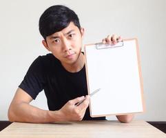 junger asiatischer mann, der ernsthaft in die kamera schaut, die ein weißes dokument in der zwischenablage zeigt und mit dem stift zeigt, sitzt am schreibtisch auf weiß isoliert, geschäftsregister-arbeitskonzept foto