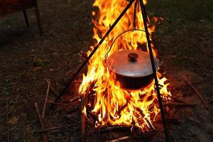 Kochen im Freien unter Feldbedingungen. Kessel auf einem Feuer im Wald. auf Reisen auf dem Scheiterhaufen kochen. Stativ mit einer Melone auf einem Feuer beim Picknick. Konzeptreisen, Trekking und Abenteuer foto