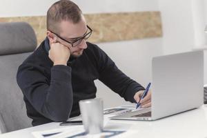 Junger Geschäftsmann, der zu Hause mit Laptop und Papieren auf dem Schreibtisch arbeitet foto