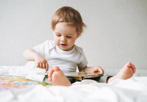 süßes lustiges Babymädchen in Hauskleidung, das zu Hause ein Buch auf dem Bett liest foto