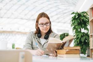 Junge brünette Teenager-Mädchen-College-Studentin mit Brille, die Hausaufgaben mit Büchern am öffentlichen Ort der grünen modernen Bibliothek macht foto