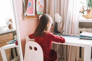 hübsches schulmädchen mit langen haaren im roten kleid zeichnet mit bleistift am tisch im kinderzimmer zu hause foto