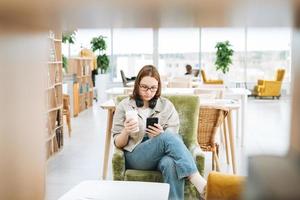 junge Brünette Teenager Mädchen College oder Gymnasiast in Brille mit Handy arbeiten in grüner moderner Bibliothek, öffentlicher Ort, grünes Freiraumbüro foto