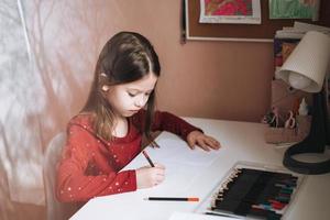 hübsches schulmädchen mit langen haaren im roten kleid zeichnet am tisch im kinderzimmer zu hause foto