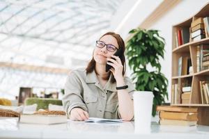 Junge brünette Teenager-Studentin in Brille mit Handy, die an einem öffentlichen Ort in der grünen modernen Bibliothek arbeitet foto