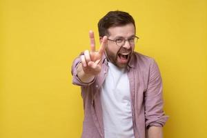 Zufriedener Mann mit Brille zeigt Siegessymbol, zwei Finger nach oben und schreit freudig. gelber Hintergrund. foto