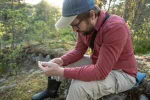 Der Mann ruht sich nach einem langen Spaziergang aus, er schaut auf sein Smartphone und sitzt auf einem mit Moos bedeckten Stein im Wald. foto