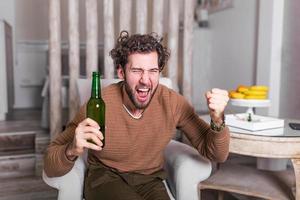 Aufgeregter Typ, der auf einer Couch sitzt, ein Bier trinkt und Fußball schaut. sport, glück und menschenkonzept - lächelnder mann, der sport im fernsehen sieht und das team zu hause unterstützt. mann, der zu hause fußball schaut foto