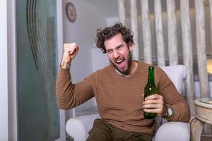 fanatischer sportfan mann, der fußballspiel im fernsehen sieht, feiert. Attraktiver, glücklicher Typ, der Fußball schaut, den Sieg seiner Lieblingsmannschaft feiert, Bier und Popcorn trinkt und im Wohnzimmer sitzt foto